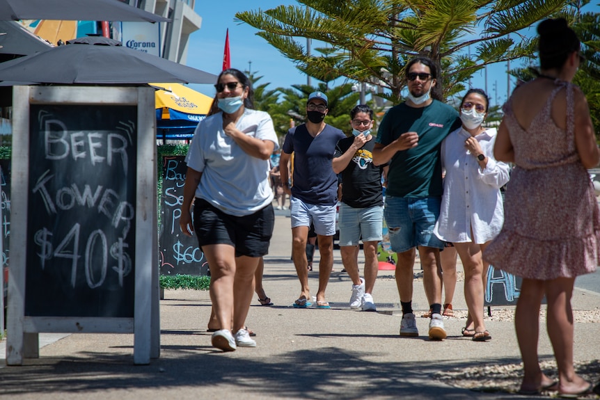 People walk along a beachside street, some wearing masks