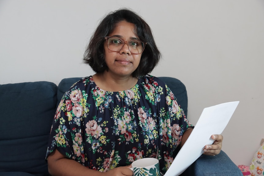 Woman with medium length dark hair and glasses smiles at camera while holding a mug and piece of paper