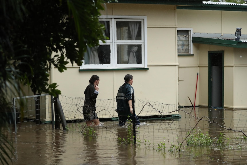 Two women walk next to a house partially submerged in floodwaters.