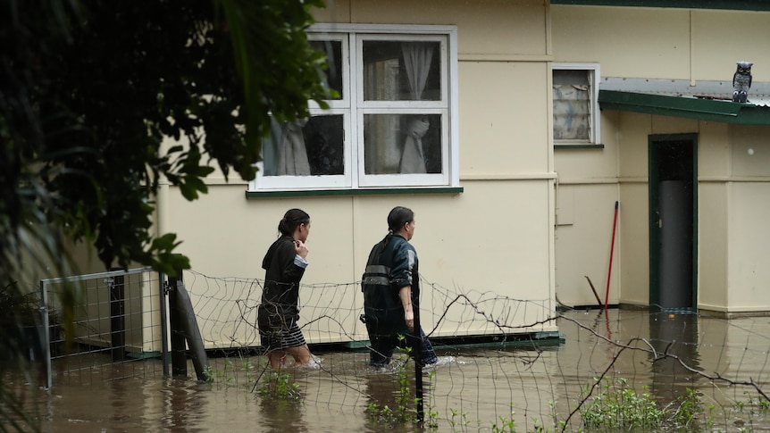 Two women walk next to a house partially submerged in floodwaters.
