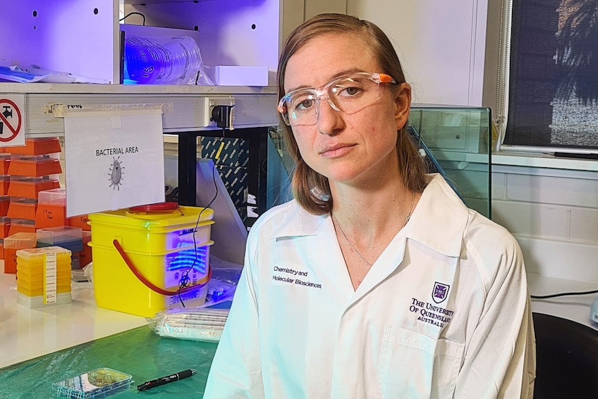 Dr Kirsty Short wearing a white lab coat and glasses, standing near a bench covered with lab items.