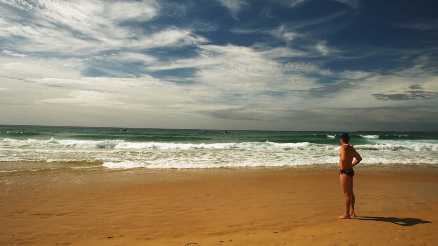 Wide shot of a man standing on a beach looking out to sea.