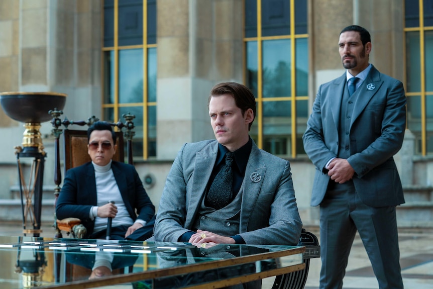 A middle-aged Asian man in a black suit and a white man in a grey suit sit at a glass table. A white man in a grey suit stands.