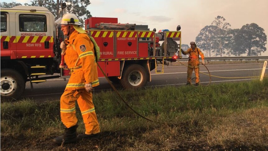 A firefighter walks on grass beside a fire truck with a hose draped over his shoulder while another pulls it from the truck.