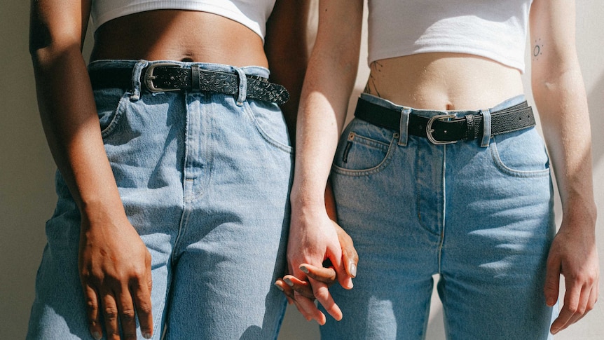 Two women holding hands, close up of their mid section and jeans in a story about the most common sex misconceptions.