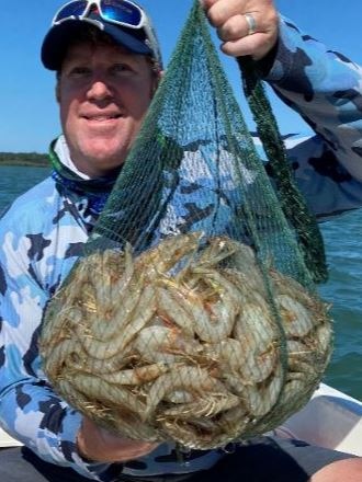 A Caucasian man in a blue fishing shirt holding up a green cast net full of prawns.