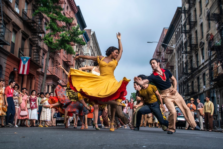 Puerto Rican woman wearing vibrant yellow dress and heels dances on street in front of crowd with Puerto Rican man in tan cinos.