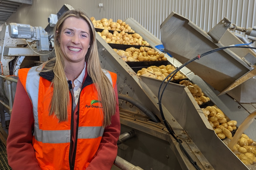 A fair-skinned blonde woman stands smiling in front of potato processing machinery.