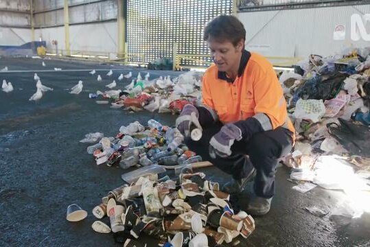 Craig Reucassel crouches beside pile of used paper takeaway coffee cups