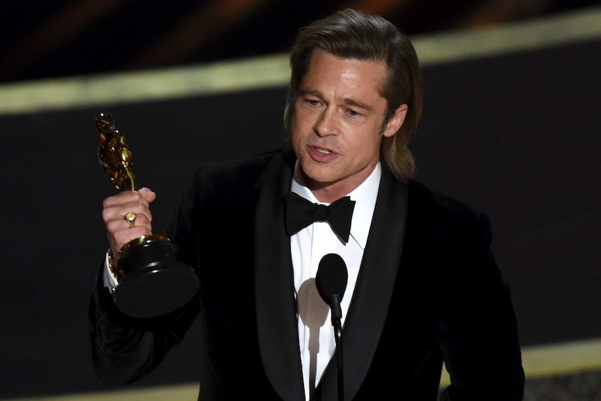 Brad Pitt, in a tuxedo, holds up a shiny gold statue