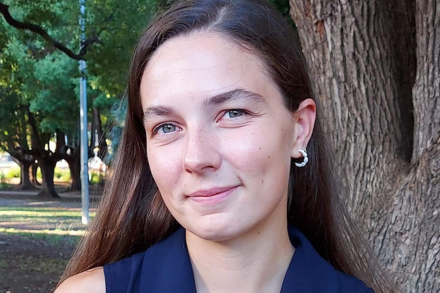 A woman with long brown hair wearing a blue shirt stands near a tree smiling.