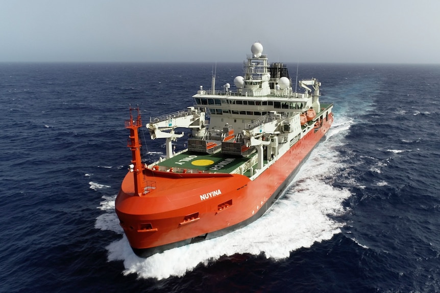 Large orange ship ploughing through dark blue ocean, with white spray washing up 
