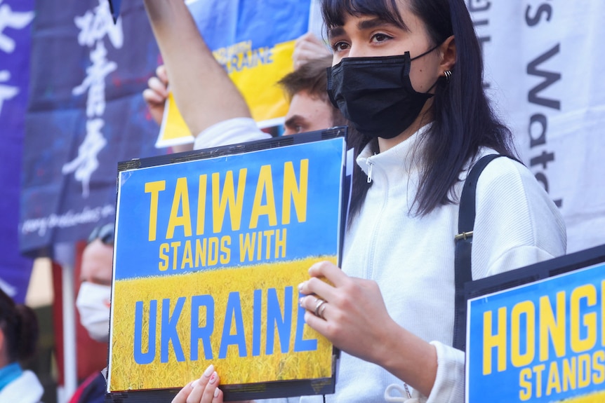 A woman wearing a blackface mask stands with a yellow and blue sign that reads TAIWAN STANDS WITH UKRAINE