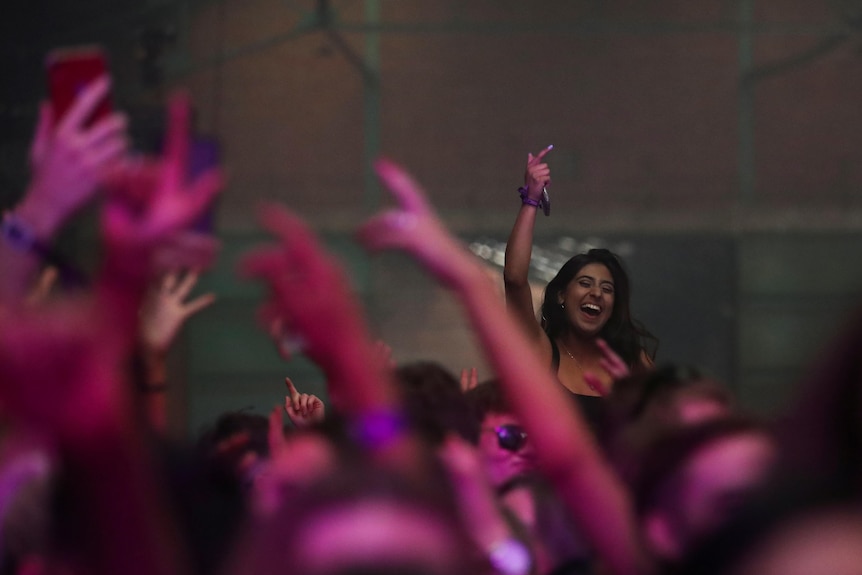 A young woman smiles as she raises her right hand while dancing among a large indoor crowd.