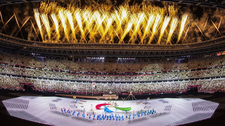 Fireworks explode into the air from the roof of a stadium filled with people as dancers dance around a big paralympic logo.