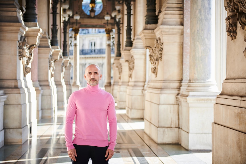 A man in a pink shirt poses for a photo in a grand hall of a European building.