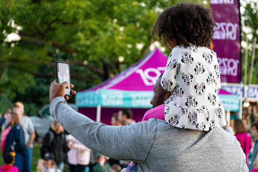 A young girl sits on a man's shoulders as he takes a selfie.
