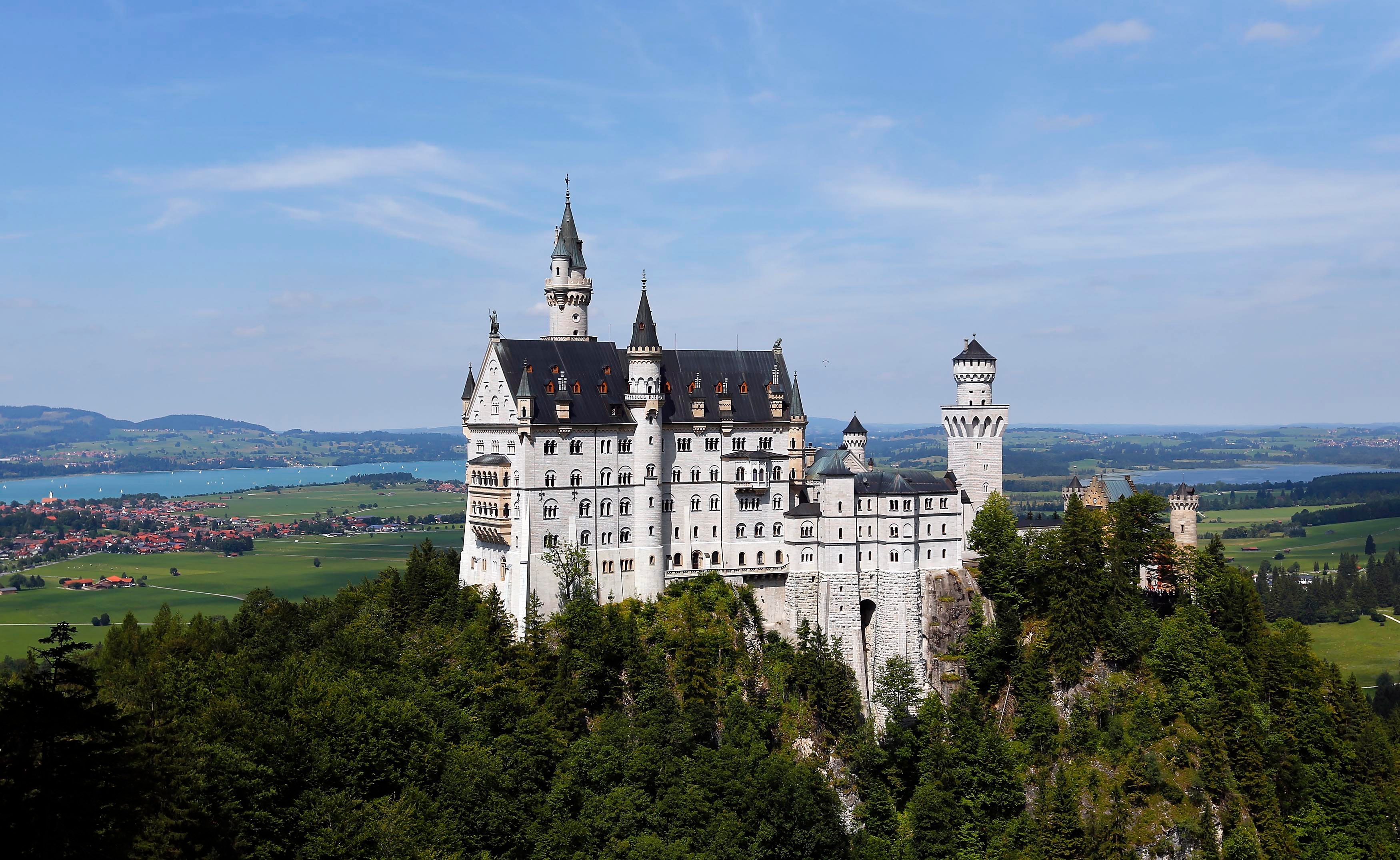 A white fairy tale-like castle is pictured in a green environment. In the background is a blue lake. 