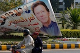 A motorcyclist passes a large sign with a picture of Imran Khan's face outside the National Assembly.