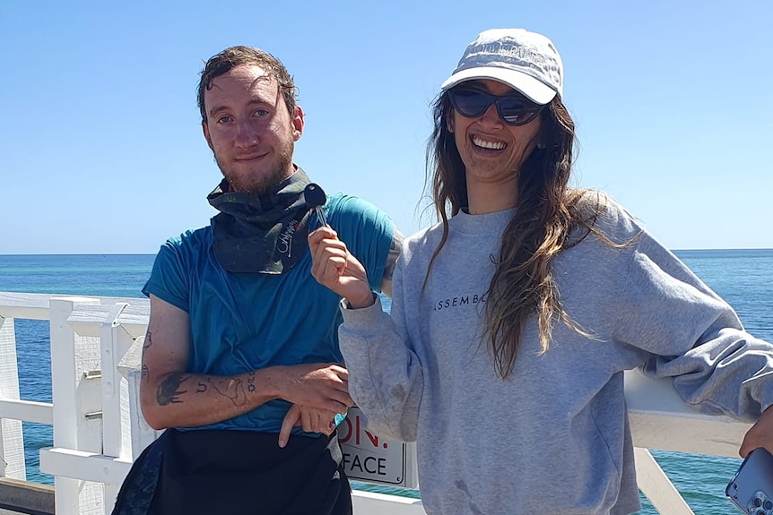 A man in tee shirt and wetsuit stands with a woman on a jetty, She is smiling holding her lost key. 