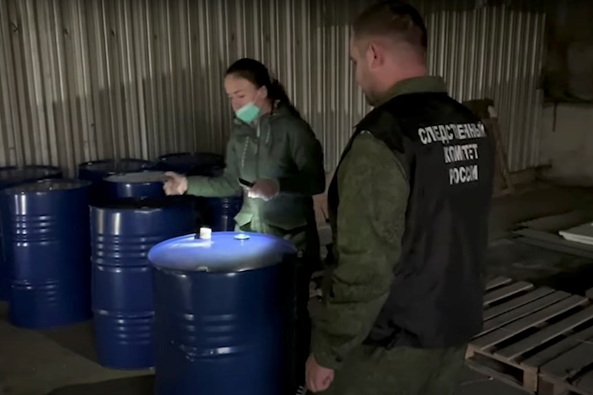 Deux personnes en uniforme de camouflage inspectent un baril bleu de 44 gallons.