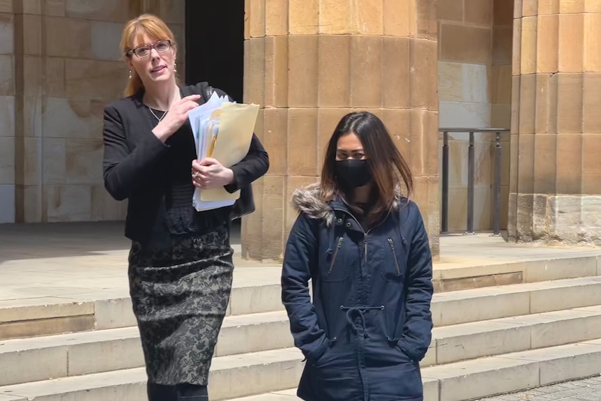 A woman wearing black and carrying a folder of paperwork walks alongside a woman with dark hair and a face mask