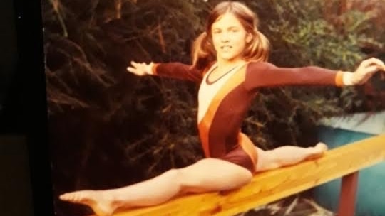 An old photo of a young girl in a leotard doing the splits on a balance beam.