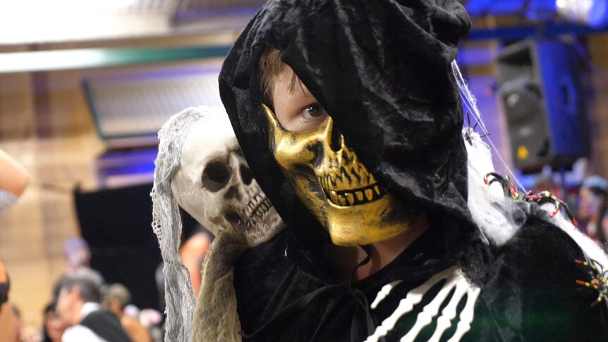 A boy dressed in a Halloween mask is cuddled by a toy skeleton