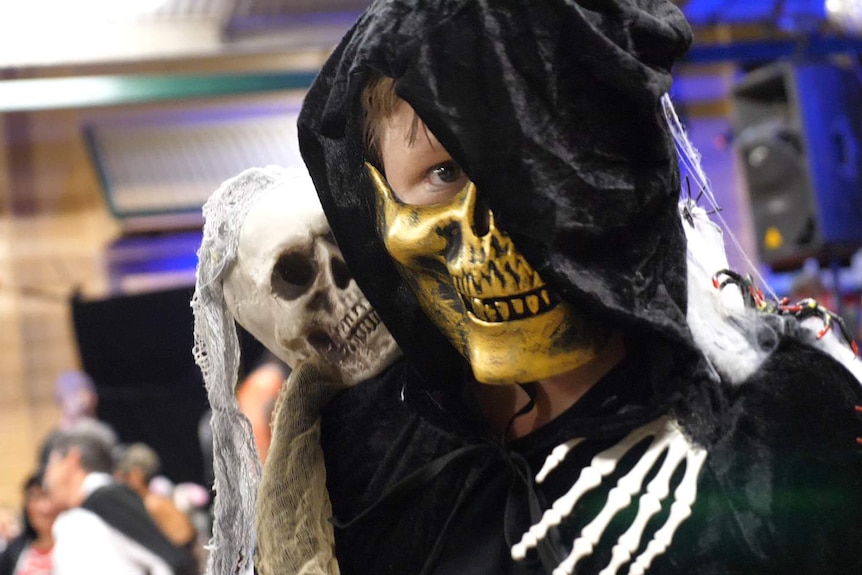 A boy dressed in a Halloween mask is cuddled by a toy skeleton