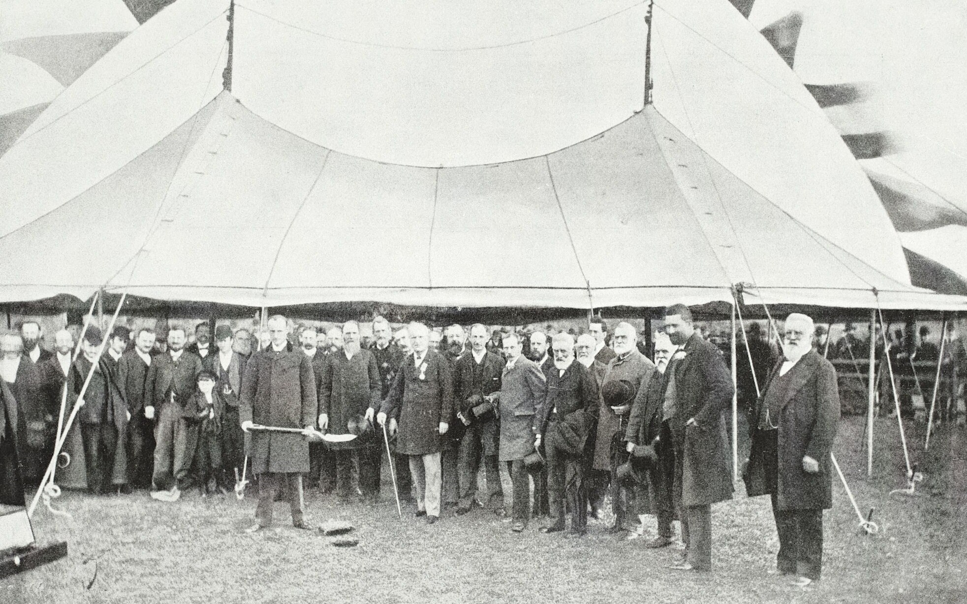 A black and white image of a crowd of people in 19th century fashion with one holding a shovel.