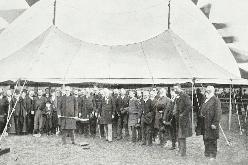 A black and white image of a crowd of people in 19th century fashion with one holding a shovel.