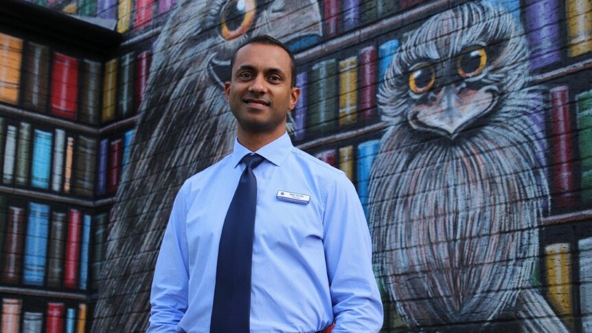 A man stands in front of a brick wall painted in a huge colourful mural of books and a giant bird