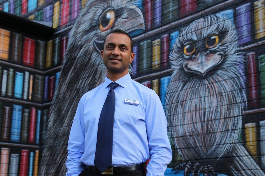 A man stands in front of a brick wall painted in a huge colourful mural of books and a giant bird