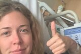 A woman with blonde hair gives a thumbs up to the camera while lying in a hospital bed.