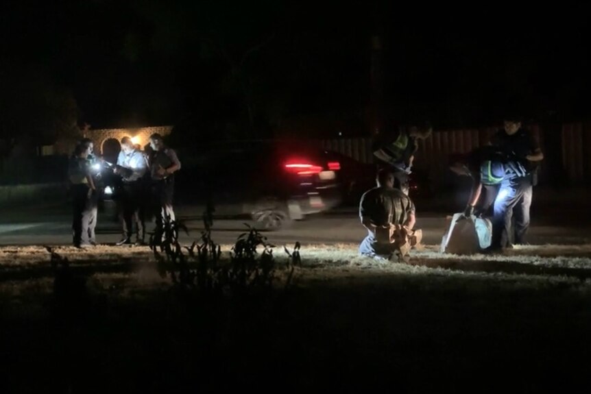 A man kneels as police arrest him outside a property, in the dark.