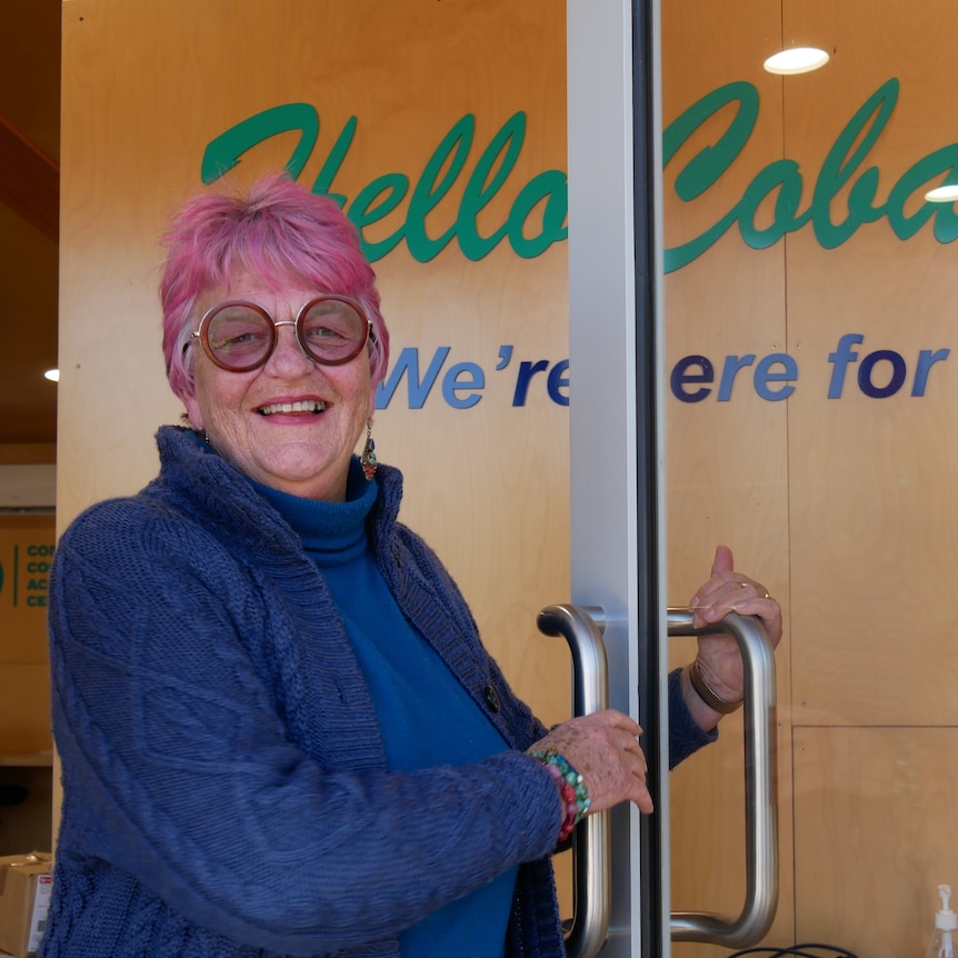 a woman smiles as she opens the door to an office space. She has pink hair and a blue jacket