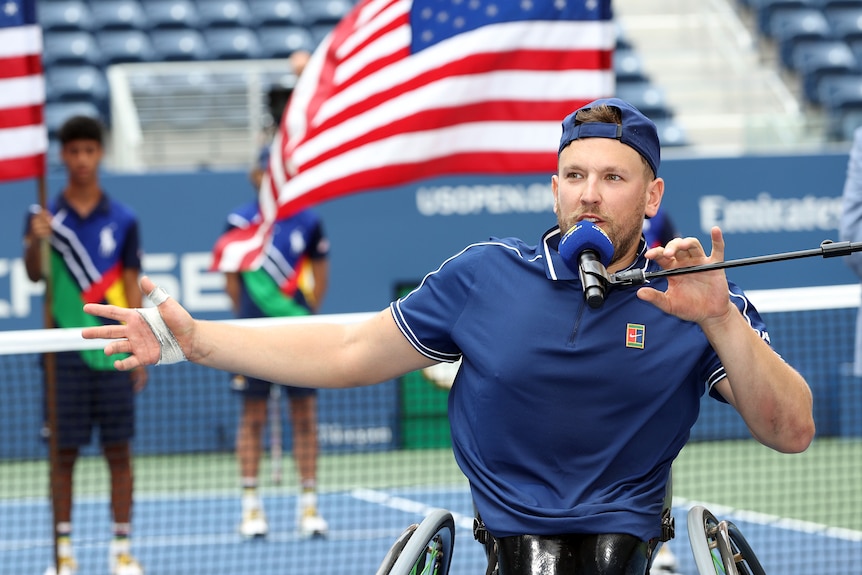 Dylan Alcott speaks into a microphone on court at Louis Armstrong Stadium, with an American flag behind him at the US Open.