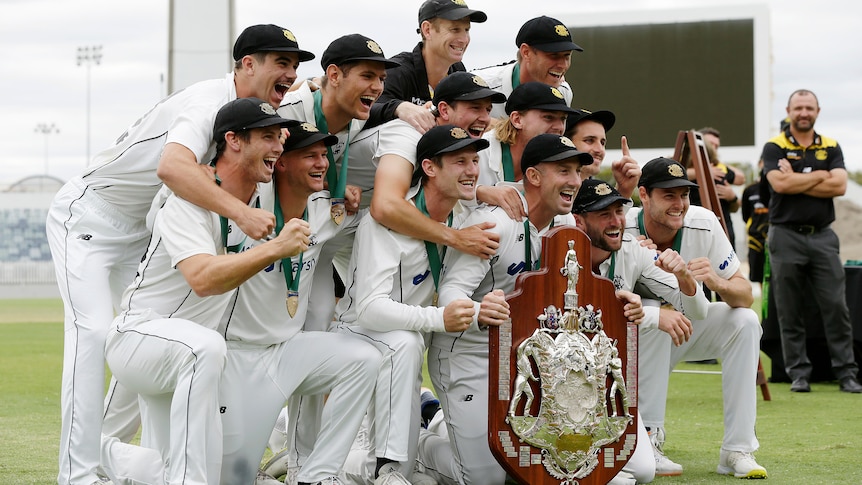 A group of men in cricket whites celebrate around a shield like trophy