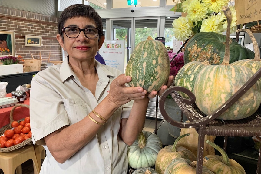 A woman holds a pumpkin alongside a display of flowers and vegetables in a cafe.