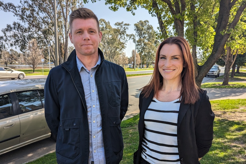 A man and a woman standing together by the side of a road, with trees in the background.