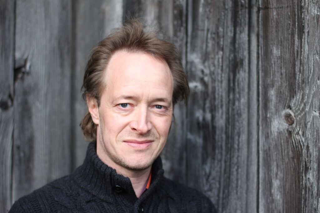 Anthony Marwood poses before a wooden fence, dressed in black and staring directly at the camera.