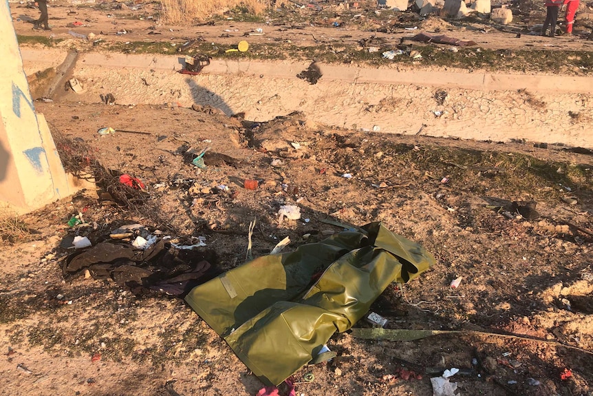 A bag and a piece of clothing along with many other items of debris lay on the ground. Two peoples' feet are seen in far right.