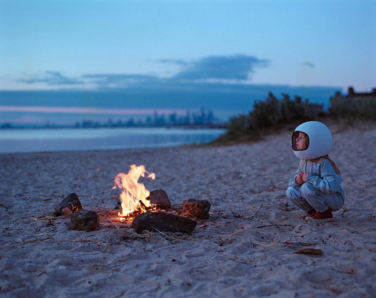 A young girl wearing a spacesuit costume and helmet crouches by a fire on the beach with the city in the background