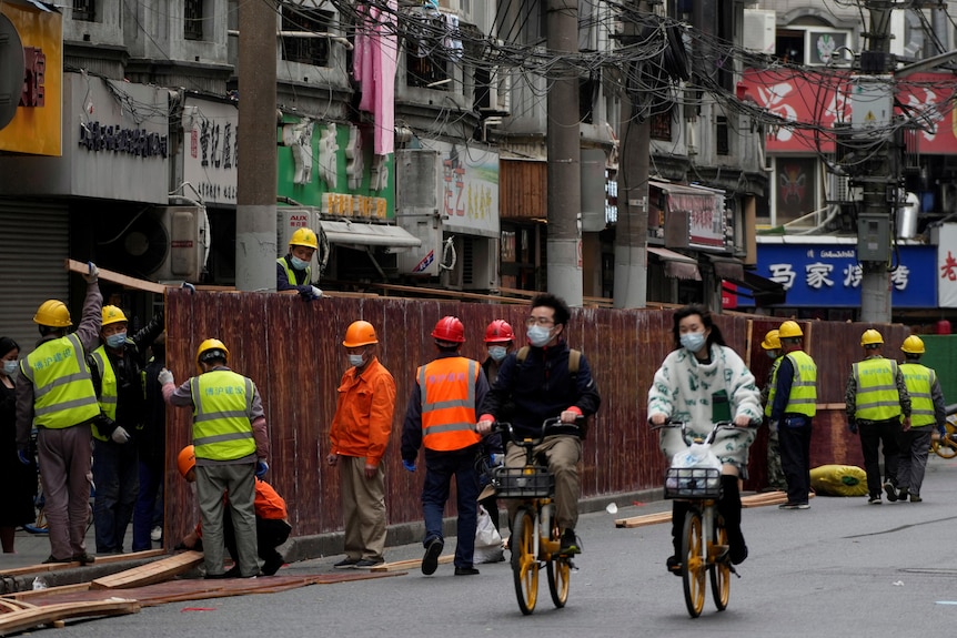 People ride bicycles past workers setting up barriers along the street.