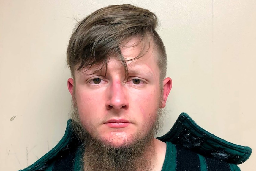 A mugshot of a Caucasian man with dark blonde hair wearing a military-style vest