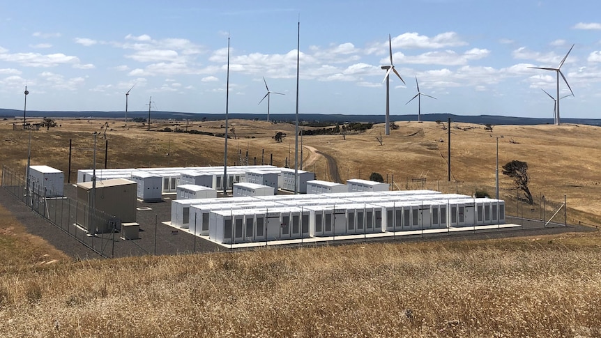 A large battery storage system sits in front of wind turbines along a hilly landscape. 