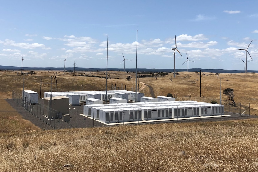 A large battery storage system sits in front of wind turbines along a hilly landscape. 