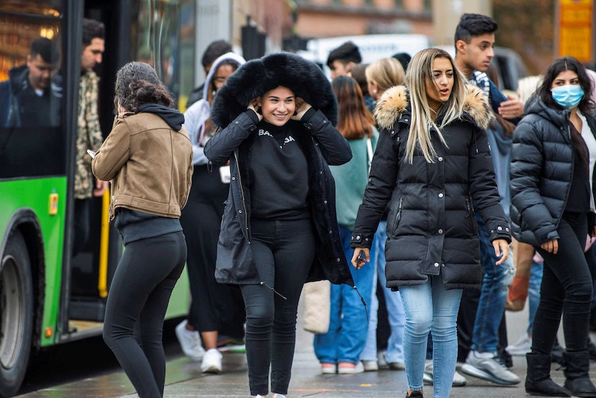 People get off a bus. Two women at the front of the crowd are walking together smiling, one wearing a thick winter coat