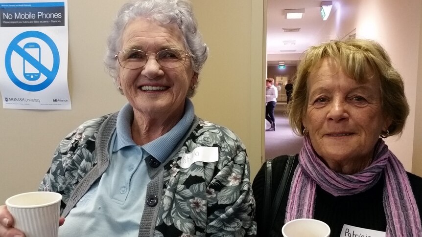 Two elderly women holding cups smile for camera.