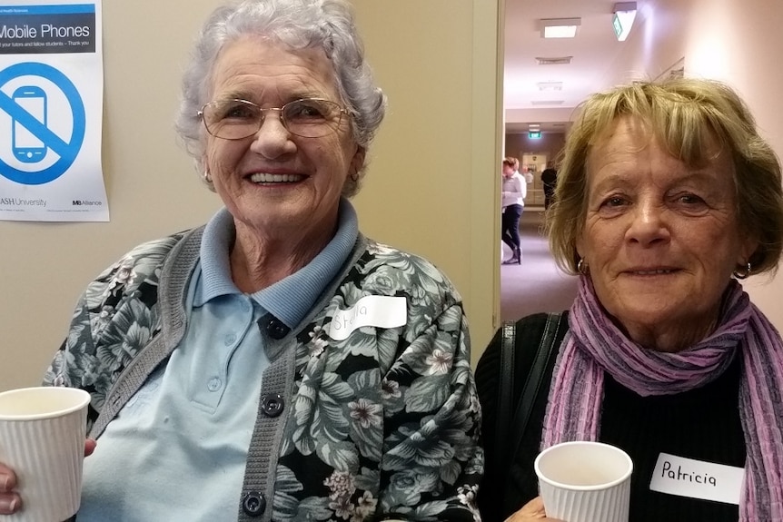 Two elderly women holding cups smile for camera.